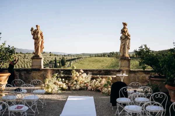 Ceremony setup for an outdoor wedding ceremony with a white aisle runner, floral ground arrangement, and chairs on a stone terrace overlook