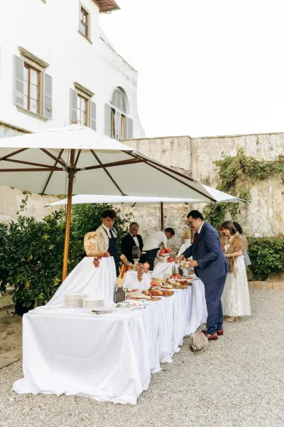 Wedding cocktail hour buffet with a cheese wheel and charcuterie on white linens under a patio umbrella in an ivy-lined villa courtyard