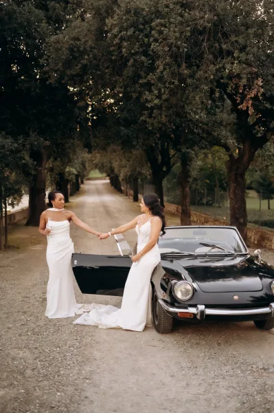 Couple portrait of two brides holding hands with champagne flutes beside a vintage convertible on a tree-lined gravel driveway