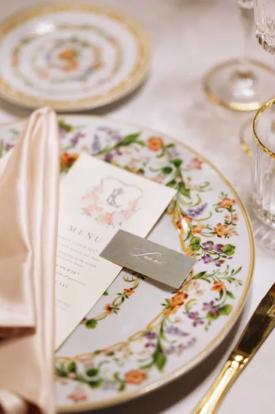 Wedding place setting with floral china plate, wedding tablescape featuring menu and calligraphy place card, blush napkin, gold flatware on white linen