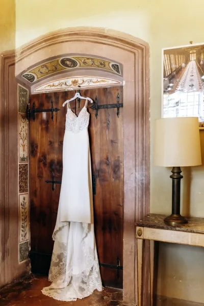 Wedding dress with lace bodice on a hanger, spaghetti straps and lace train framed by rustic wooden doors and arched murals indoors
