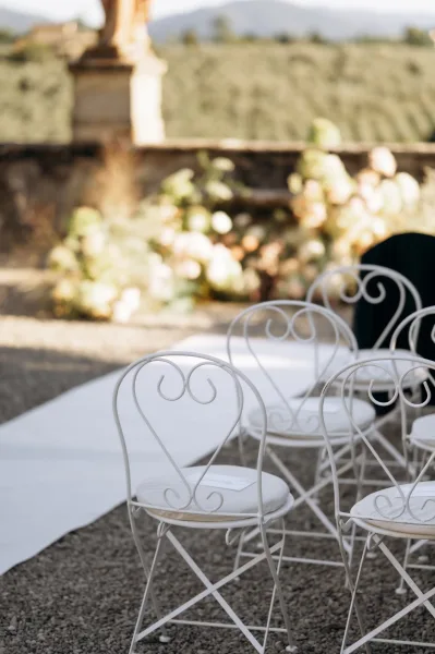 Ceremony aisle decor with a white aisle runner flanked by white metal chairs and floral arrangements, set on gravel by a stone wall and mountains