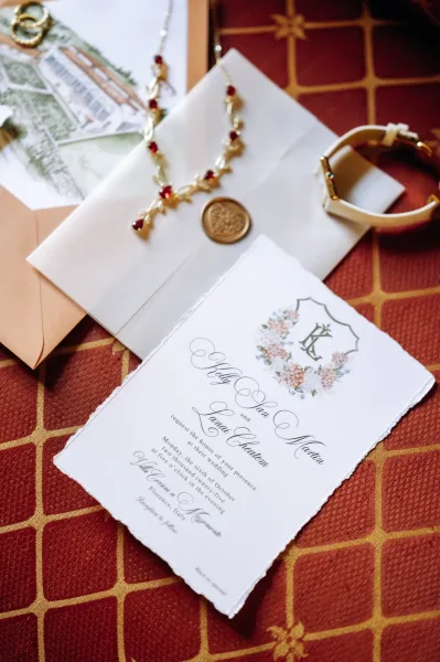 Invitation suite flat lay with deckle edge wedding invitation, vellum wrap, wax seal, rings and gold jewelry on patterned red fabric