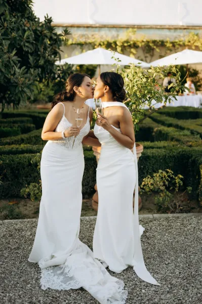 Wedding kiss portrait of two brides kissing while holding champagne flutes on a gravel garden path with hedges and patio umbrellas