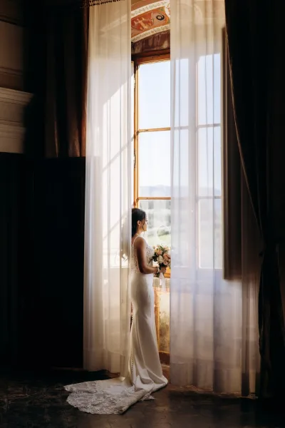 Bridal portrait of a bride by window in a fitted lace gown with long train, holding a rose bouquet with ribbon in soft natural light