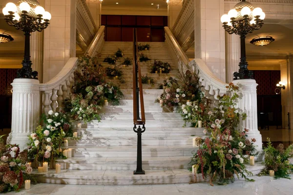Wedding staircase decor with staircase floral installation, greenery garlands and pillar candles on a marble staircase under globe chandeliers in a grand lobby