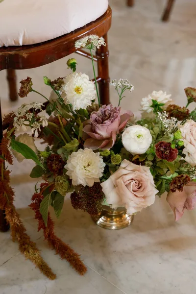 Wedding bouquet with roses and dahlias in a brass vase on a marble floor beside a wooden chair with a white cushion