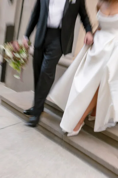 Wedding recessional as bride and groom walking down stone steps, bride lifting her gown with bouquet and heels, groom in black tuxedo