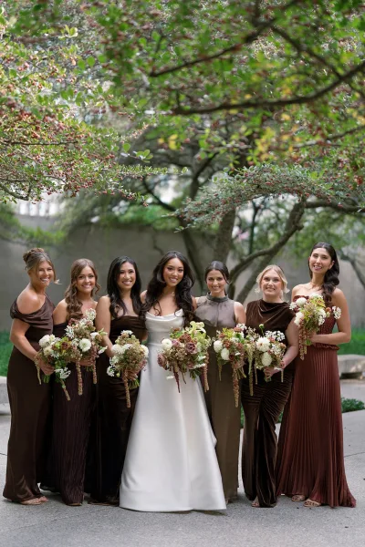 Bridesmaid group photo with bride in a white gown and bridesmaids holding bouquets, standing by a stone wall in a garden walkway