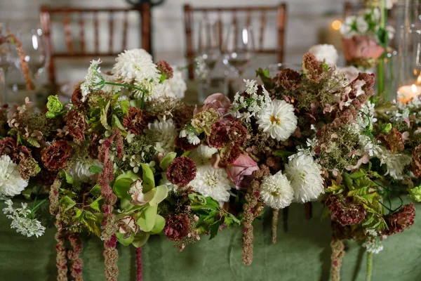 Wedding table centerpiece with a floral table runner of dahlias, orchids, and greenery, accented by taper candles and wine glasses in a reception room