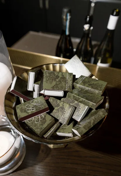 Wedding matchboxes with custom wedding matchboxes monogram sleeves arranged on a brass tray beside candles, glass hurricane holders, and wine bottles