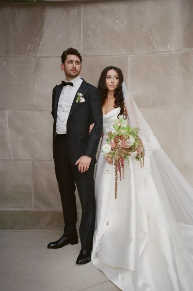 Couple portrait of bride and groom, bride holding bouquet on his arm in strapless dress and long veil, groom in black tux by stone wall