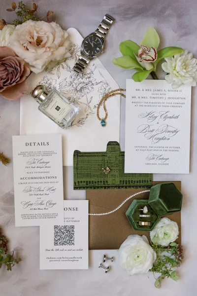 Wedding invitation suite flat lay with letterpress wedding invitation, RSVP and details cards, rings, watch, perfume, and floral liner on linen backdrop
