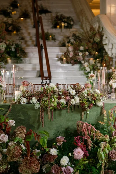 Reception tablescape with head table flowers, lush floral runner, greenery garland, and taper candles on stone steps by a grand staircase
