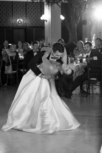 First dance dip as groom in tuxedo dips bride in satin ball gown for a kiss on ballroom dance floor, guests watching near candlelit tables