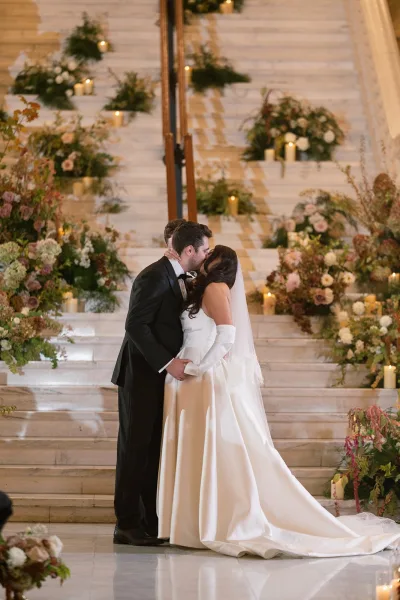 Wedding kiss portrait of bride and groom kiss on a marble staircase, her long veil and opera gloves framed by candles and lush florals