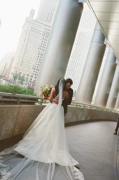 Couple portrait of bride and groom embrace on an urban walkway, her long train and veil flowing as she holds a bouquet by modern columns