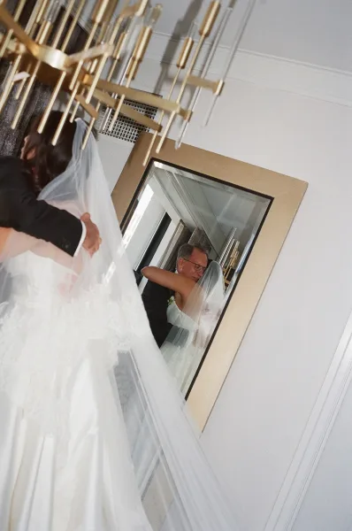 First look moment as bride in strapless gown and veil hugs her father beneath a chandelier in a white-paneled hallway by a mirror