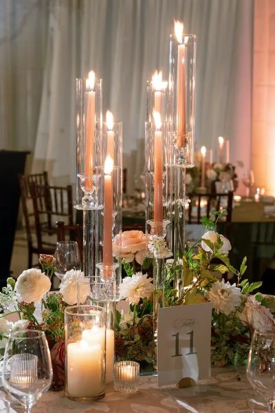 Reception tablescape with wedding candle centerpiece of taper and pillar candles in glass holders, white florals and greenery under warm uplighting