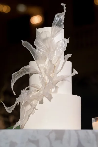 Wedding cake with translucent sugar sail decor on a white tiered wedding cake beside a votive candle in a dim reception room with warm bokeh lights
