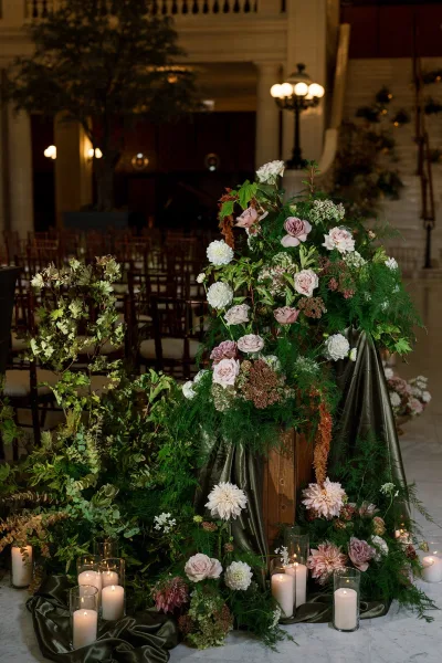 Ceremony aisle flowers in a wedding aisle floral arrangement with blush roses, white dahlias, and eucalyptus on a wood pedestal beside candles indoors