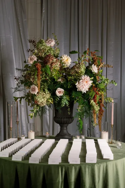 Escort card display with wedding escort cards beside a black urn floral centerpiece, taper and votive candles on a green tablecloth before gray drapery