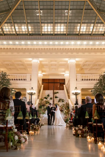 Ceremony moment at an indoor wedding ceremony with bride and groom beneath a floral arch, candles lining the aisle before a grand staircase hall