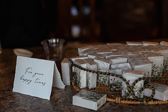 Wedding favors in floral printed wrappers arranged in a woven basket tray with favor boxes and a place card on a stone countertop