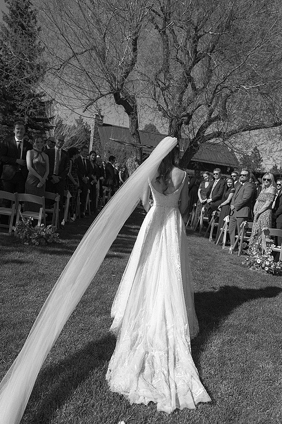 Wedding processional with bride walking down aisle in lace dress and cathedral veil, guests seated along floral-lined lawn aisle by trees