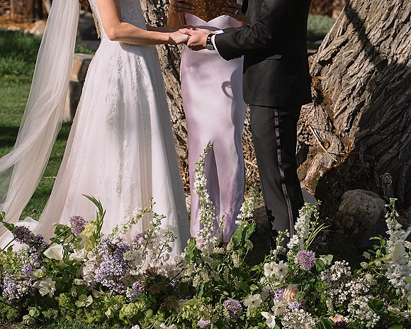 Wedding vows as bride and groom holding hands, lace dress and veil beside tuxedo, with purple and white aisle flowers on a garden lawn
