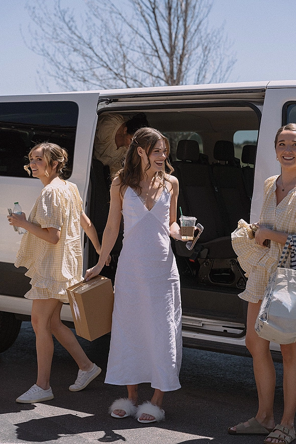 Getting ready moment as bride steps out of a van in a white slip dress with hair clips, holding iced coffee in a parking lot under blue sky
