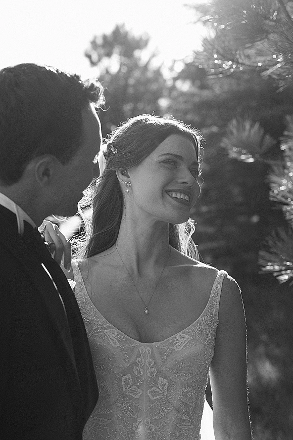 Wedding couple portrait in black and white, bride in lace dress and veil smiling over shoulder as groom in tux embraces her outdoors in sunlight