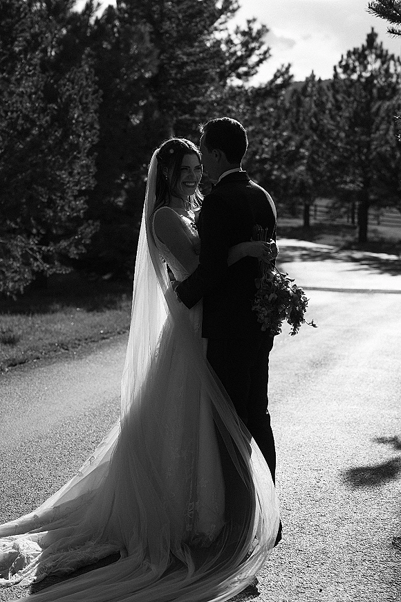 Couple portrait of bride and groom embrace, her long veil and bouquet flowing as they hug on a sunlit, tree-lined road