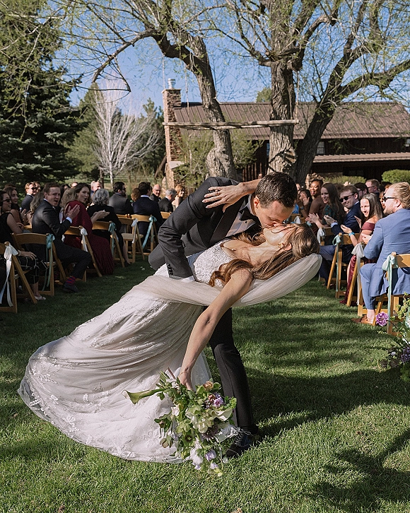 Wedding kiss portrait of bride and groom in a dip kiss wedding photo, veil flowing, holding a greenery bouquet on a sunny outdoor aisle with guests behind