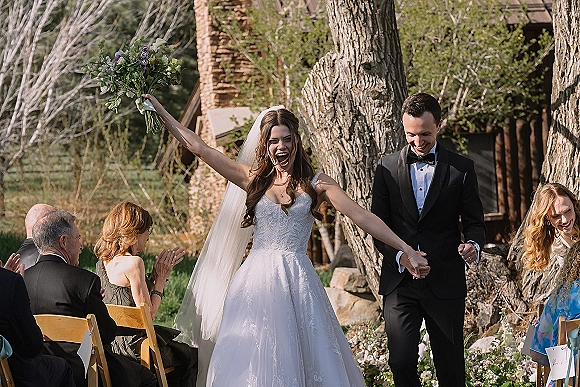 Wedding recessional as newlyweds walk hand in hand down the aisle, bride raising her bouquet, guests clapping amid trees and rustic stone venue