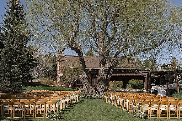 Ceremony setup for an outdoor wedding ceremony with wood folding chairs lining a grassy aisle, florals and ribbons near a rustic lodge backdrop