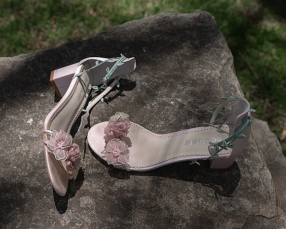 Wedding shoes with floral applique accents, ankle ties, and block heels resting on stone rock beside a grass lawn