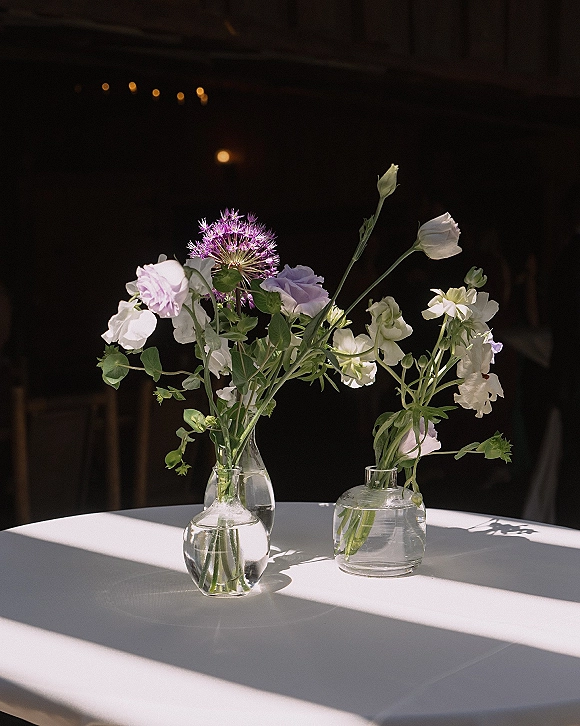 Wedding centerpieces with bud vase centerpiece in clear glass vases, white and lavender flowers with greenery on linen tables under bistro lights