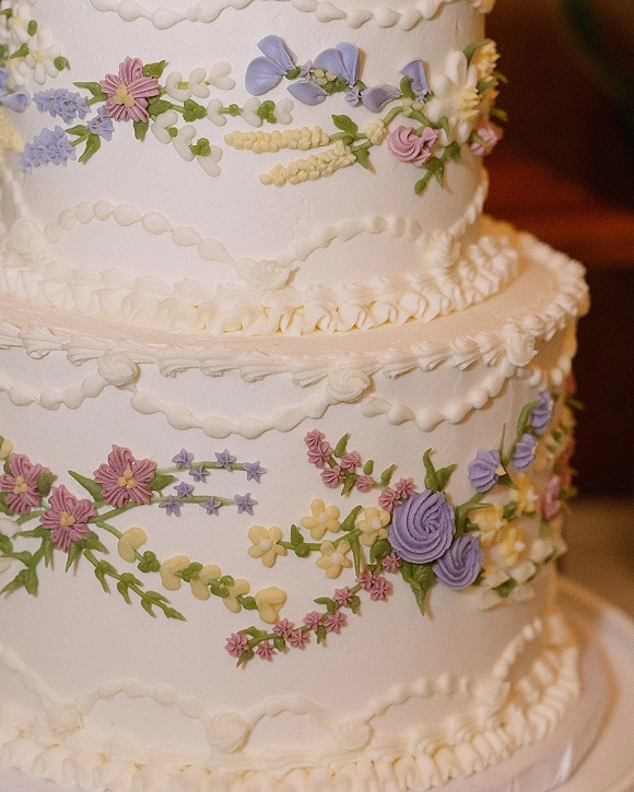 Wedding cake with buttercream wedding cake ruffle piping and pastel floral garland, shown in warm indoor reception lighting with blurred background