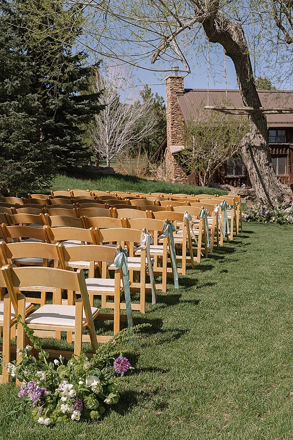 Outdoor ceremony seating with wedding ceremony chair rows of wood chairs, ribbon ties, and aisle florals on a lawn by a cabin under trees