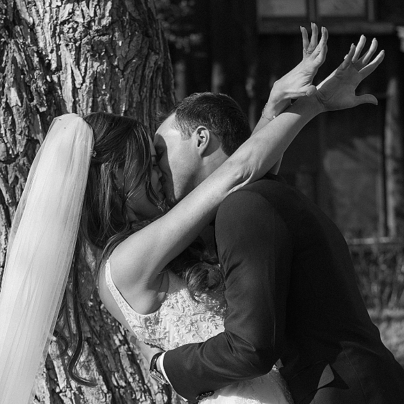Wedding kiss in a black and white wedding photo as bride and groom embrace by a tree trunk, her veil and lace dress visible