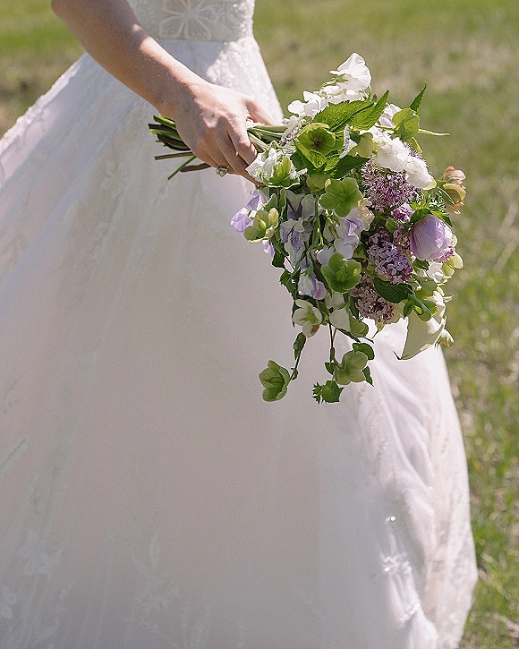 Bridal bouquet with white and purple flowers and trailing greenery held against a lace bodice, wedding ring visible on an outdoor lawn