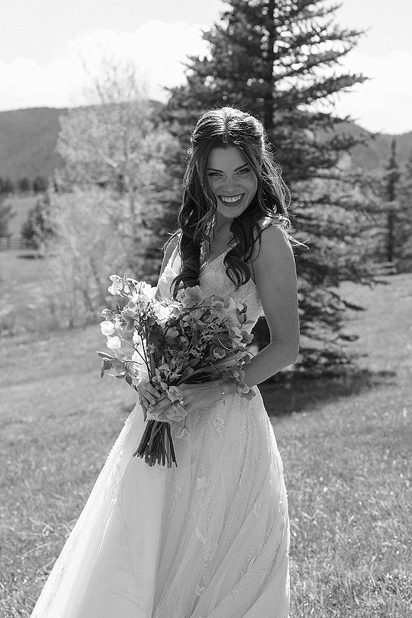 Bridal portrait of a smiling bride holding a bouquet in her lace wedding dress, posed in a meadow with mountains and evergreens behind