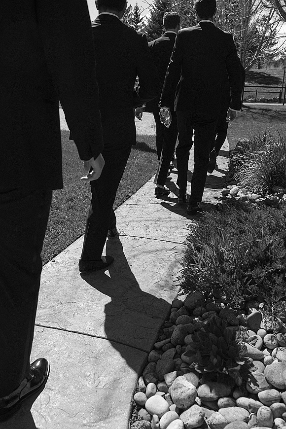 Groomsmen portrait of men in black suits walking away on a stone walkway, dress shoes aligned, one holding a water bottle near shrubs and trees