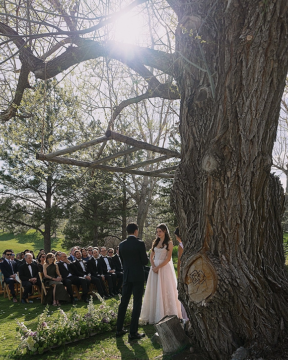 Wedding ceremony under a large tree with bride in veil and groom in black tuxedo exchanging vows beside floral aisle arrangements in sunlit garden