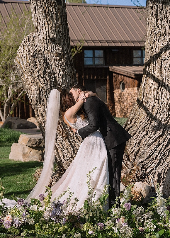 Wedding kiss portrait of bride and groom kissing, her lace dress and veil flowing as he holds her waist on a grassy lawn by a rustic building