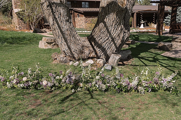 Wedding ceremony florals with grounded ceremony flowers in roses, lilac, and white blossoms arranged along a garden walkway near a pergola