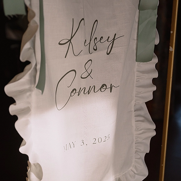 Wedding welcome sign on white fabric with calligraphy lettering, ruffled trim, sage green ribbon accent, and gold frame indoors