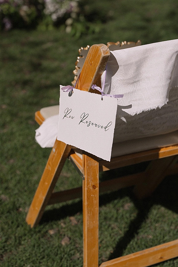 Wedding reserved seat sign with calligraphy text on a paper tag tied by lavender ribbon to a wooden chair with white cover on grass lawn