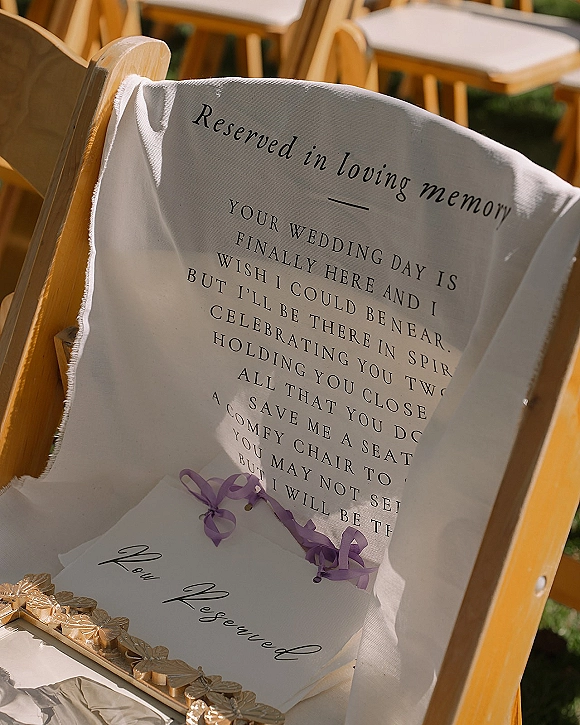 Reserved seat sign on a wooden wedding memorial chair draped in white fabric with a lavender ribbon, set among outdoor ceremony chairs on grass lawn